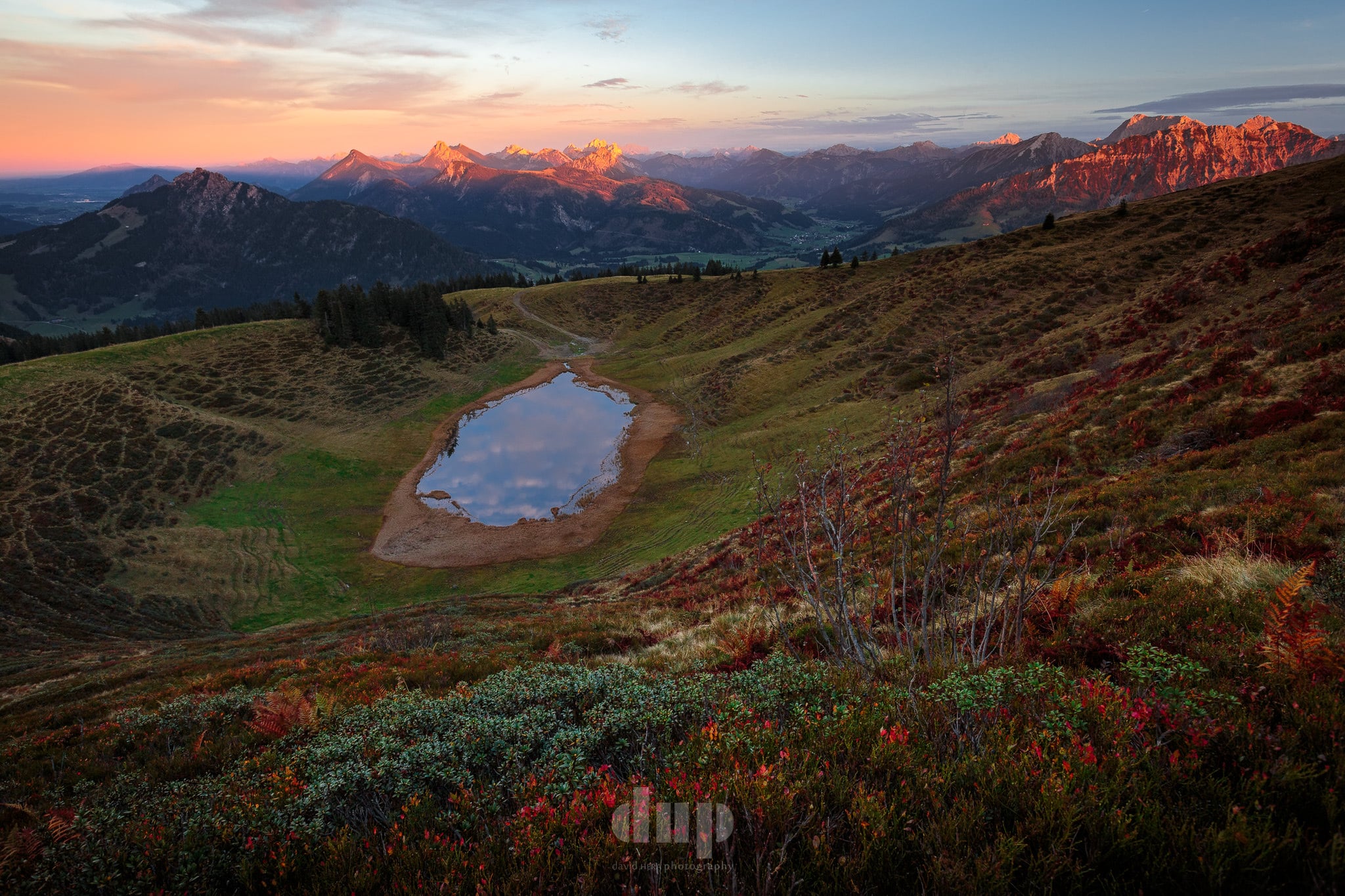Alpine tarn at sunset, golden mountains in the distance, autumnal foliage in the foreground.